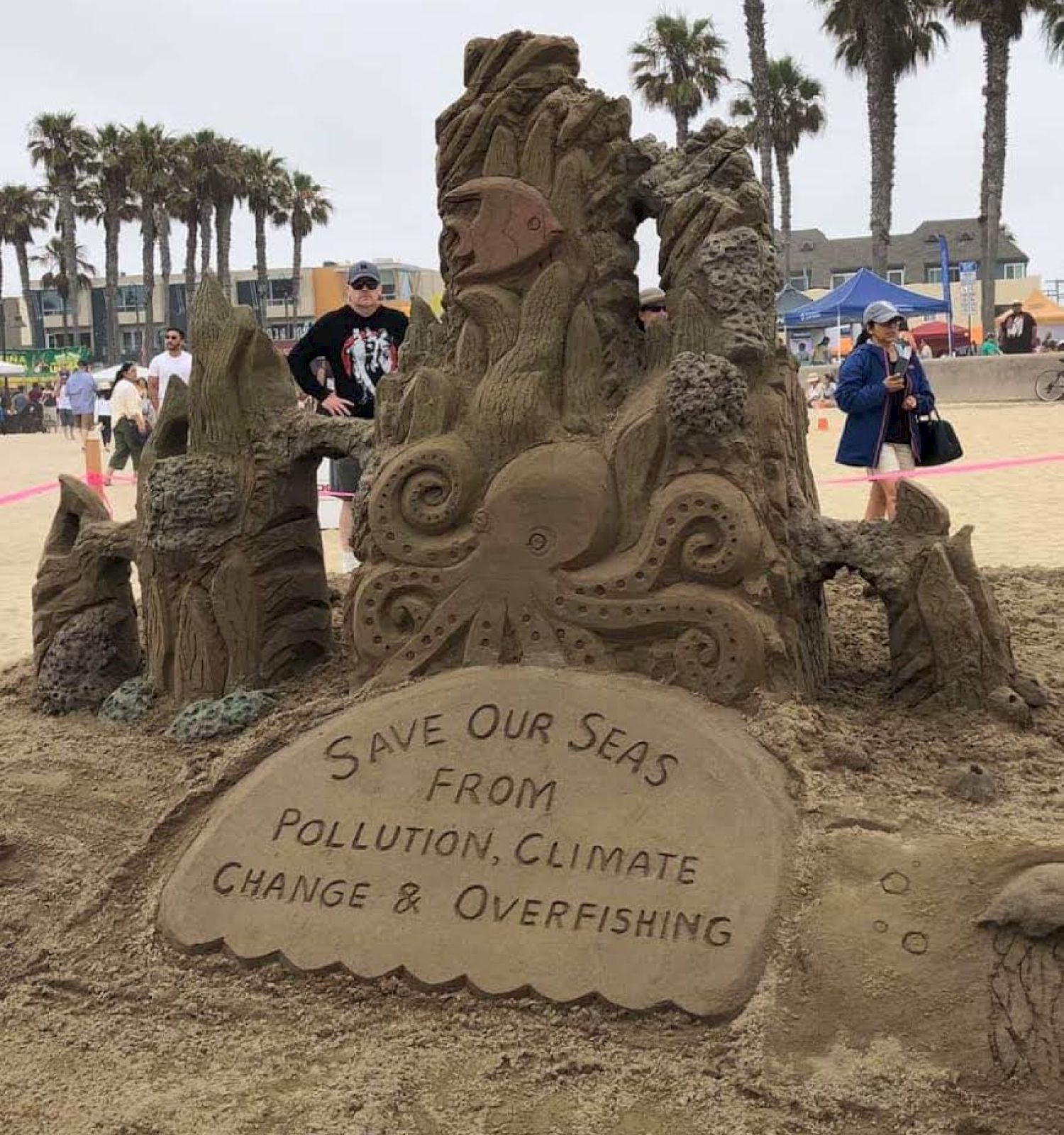 A sand sculpture at a beach with a message: "SAVE OUR SEAS FROM POLLUTION, CLIMATE CHANGE & OVERFISHING." Palm trees and people in the background.