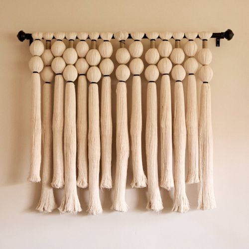 The image features a decorative wall hanging made of suspended, textured beige strands and large beads, creating a visually pleasing pattern.