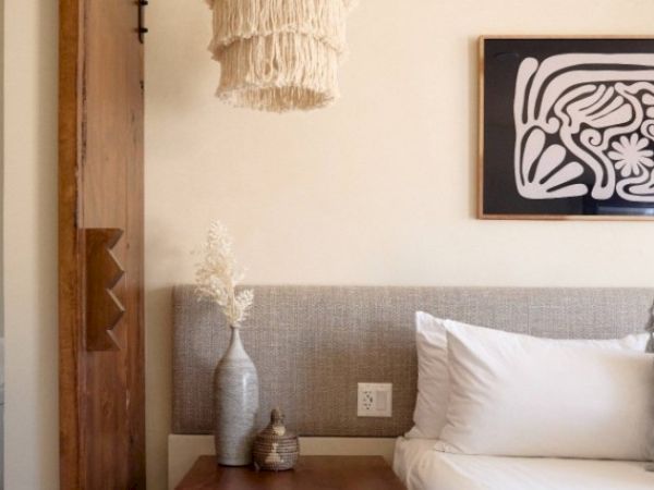 A cozy bedroom with a headboard and a nightstand holding a vase and a small decorative item, a framed abstract artwork, and a hanging woven light.