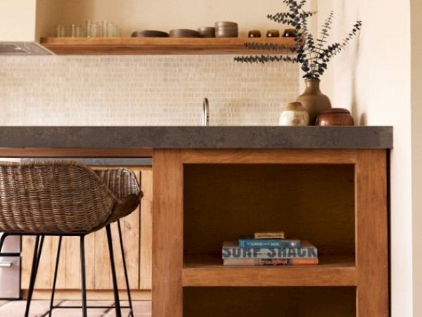 A cozy kitchen features wooden shelves, a gray countertop, wicker stools, various pottery, and a cookbook titled "A Surf Shack" on the lower shelf.