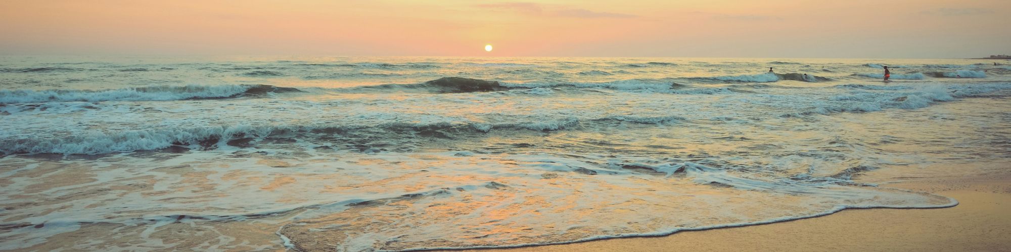 A serene beach at sunset with gentle waves lapping against the shore and a colorful sky with clouds illuminated by the setting sun.