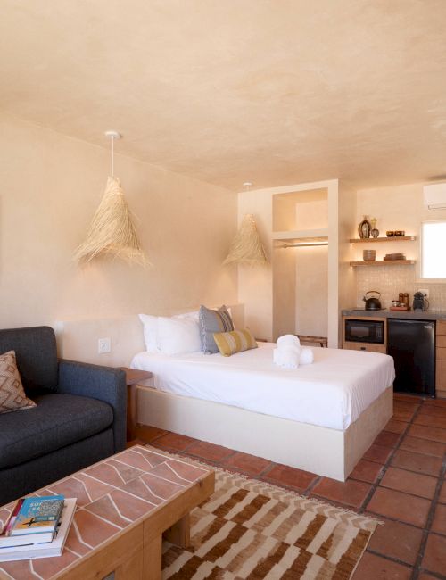 A cozy room features a bed, sofa, wooden table, kitchenette, and wall art, with warm lighting and tiled flooring.