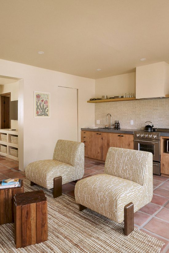 The image shows a cozy living area with chairs, a sofa, a small kitchen with appliances, and a visible bedroom, all in neutral tones.